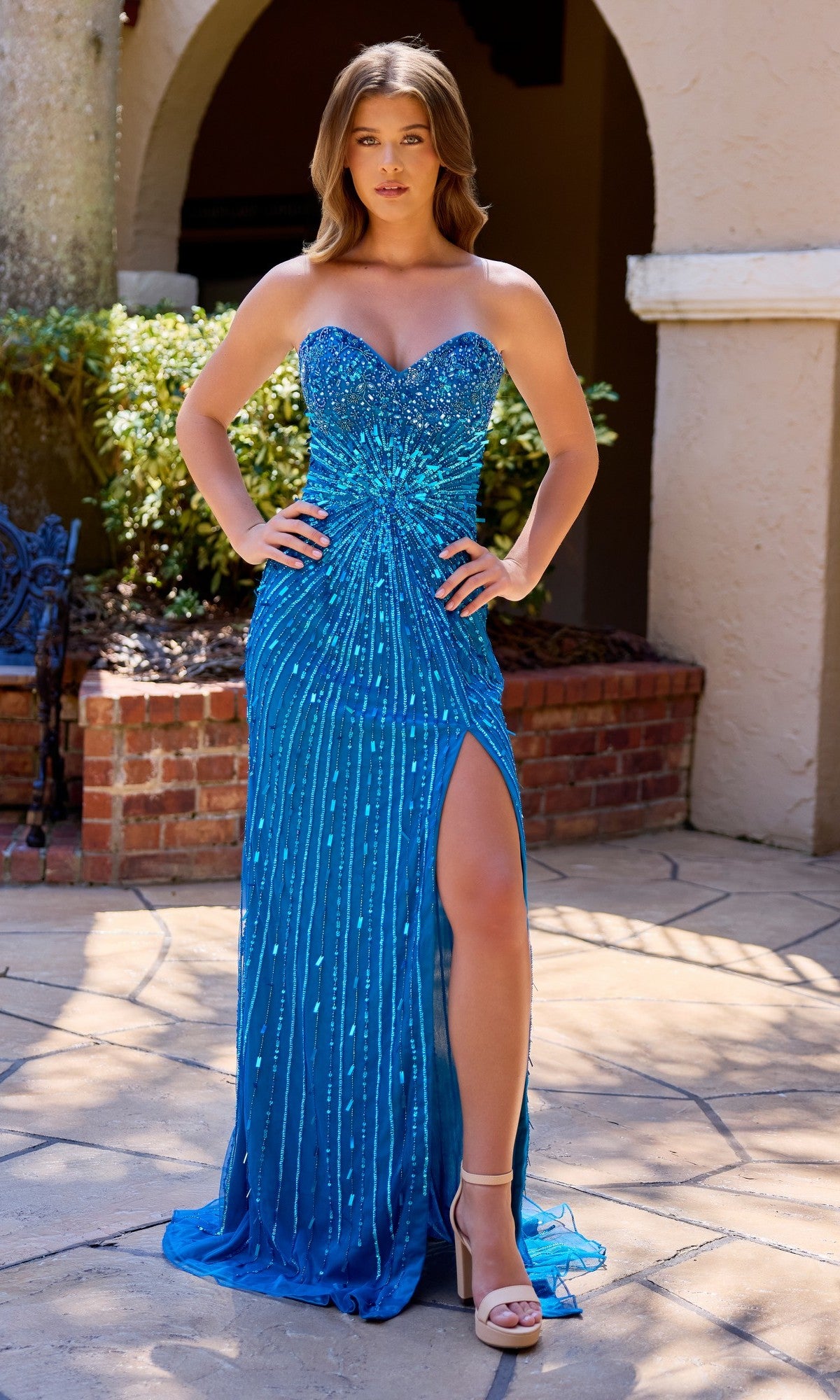 A woman poses outdoors in the Primavera 4512 long prom dress in Peacock, featuring a strapless beaded bodice, star-bust pattern, and high-leg slit. She pairs it with high-heeled sandals; her long hair flows against a backdrop of shrubs and brick wall.